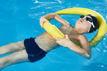 Preschool boy learning to swim in pool with foam noodle