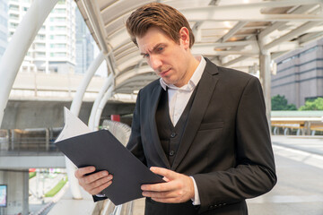 Fototapeta premium Businessman in a black suit reading documents outdoors in a modern city background. Concept of career, corporate work, planning, decision-making, and professional lifestyle.