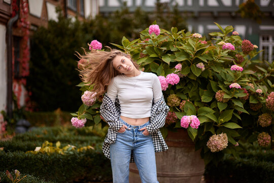 Young woman in casual autumn outfit posing among blooming hydrangeas in front of traditional half-timbered house in Germany &ndash; cozy and stylish outdoor portrait with warm fall colors