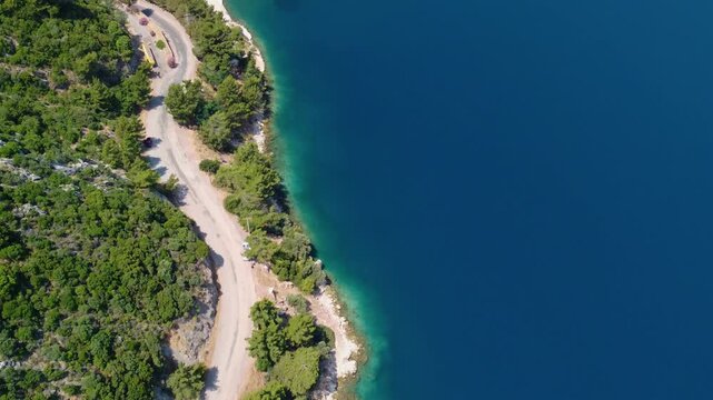 Drone footage captures a scenic aerial view of a winding coastal road nestled amidst lush green hills, bordering the vibrant turquoise waters of the Mediterranean Sea near Icmeler, Turkey.