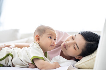 Loving asian mom carying of her newborn baby at home. Mother and baby boy playing in sunny bedroom. Parent and little kid relaxing at home. Family having fun together. Childcare, maternity concept.