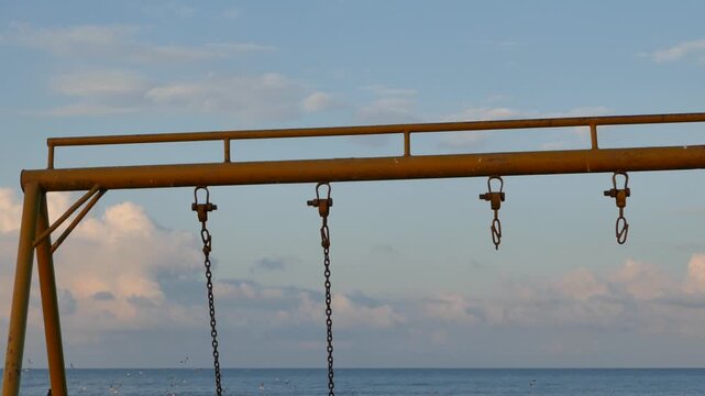 Close-up of an old swing set structure with rusty chains hanging under a cloudy sky. A nostalgic, symbolic view of childhood memories, time passing, and the contrast between play and decay.