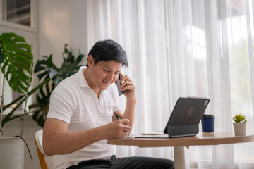 Asian freelancer talking on phone and taking notes in home office