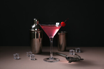 Glass of tasty Cosmopolitan cocktail garnished with cherry and ice cubes on brown table against black background