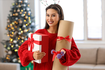Young woman with gift box and fitness mat at home on Christmas eve