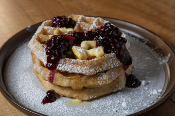 Close-up of golden waffles topped with butter, fresh huckleberries, and berry jam as powdered sugar is sprinkled on top. Rustic breakfast or dessert food photography with a gourmet, homemade style.