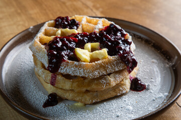 Close-up of golden waffles topped with butter, fresh huckleberries, and berry jam as powdered sugar is sprinkled on top. Rustic breakfast or dessert food photography with a gourmet, homemade style.