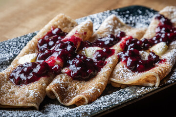 Close-up of homemade crepes topped with fresh huckleberries, berry jam, and powdered sugar on a rustic wooden table.
