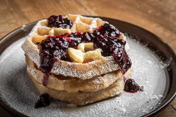 Close-up of golden waffles topped with butter, fresh huckleberries, and berry jam as powdered sugar is sprinkled on top. Rustic breakfast or dessert food photography with a gourmet, homemade style.