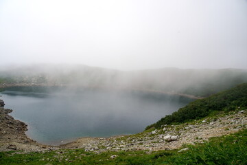 霧に覆われた高山にひっそりと佇むカルデラ湖または火口湖を捉えた風景写真湖面は静かに深く青い緑の草木と岩場のコントラストが印象的