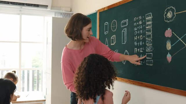 Skilled caucasian teacher looking at african girl writing answer on blackboard at elementary school. Professional student walking at in front of class to answer quiz in elementary classroom. Pedagogy.