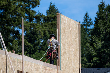 New housing development construction project, carpenter on wall in safety harness and hardhat, using pneumatic nail gun, work in progress

