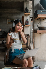 Woman in casual attire with headphones, holding coffee, working on a laptop in a stylish industrial cafe setting.