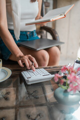 A woman calculates expenses while reading, surrounded by flowers and coffee, creating a cozy and productive atmosphere.