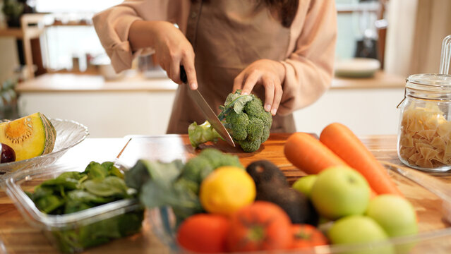 A woman is cutting a broccoli on a wooden cutting board. The kitchen is well-stocked with various fruits and vegetables, including apples, carrots