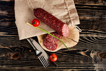 A piece of fresh smoked sausage, cutlery and cherry tomatoes in close-up as decoration on a dark wood background.