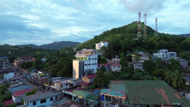 Aerial view toward Mount Tapyas in Coron town with hillside telecom towers colorful buildings roadside shops and tropical greenery under cloudy light
