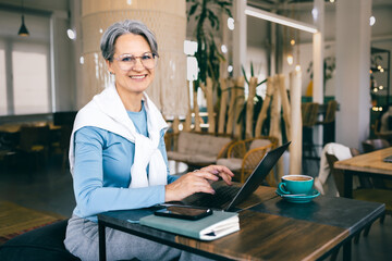 Smiling senior freelance woman with gray hair working on laptop in cafe, confident and independent