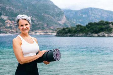 Smiling senior woman holding yoga mat on beach, embracing active and healthy aging outdoors