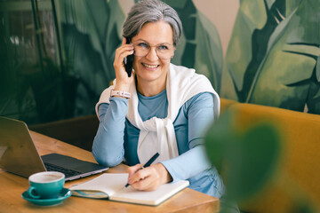 Senior woman in casual outfit taking notes and having a phone call at cozy freelance workspace