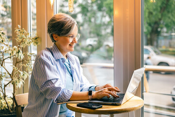 Senior woman smiling while working on laptop at cozy cafe table by the window with natural daylight