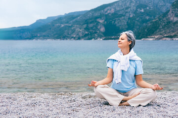 Senior woman meditating by the sea, enjoying peaceful moment and mountain coastal scenery