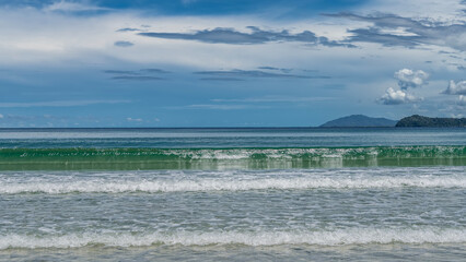 The waves of the turquoise ocean roll in rows to the shore, foaming. Reflection. Mountains on the horizon. Blue sky, clouds. A sunny summer day. Malaysia. Borneo. Kota Kinabalu.
