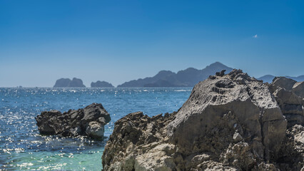 Picturesque cliffs in the ocean. Reflections on the surface of the turquoise sea. Mountains on the horizon. In the foreground is a rock with rough furrowed slopes. Clear blue sky. Copy space.