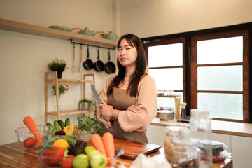 A woman is standing in front of a table with a variety of vegetables and fruits, including carrots, tomatoes, and broccoli. She is holding a knife and she is preparing a meal