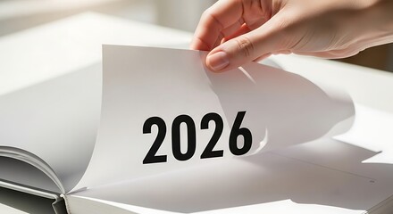 A hand turns a page in a book, revealing the year 2026, symbolizing a new beginning or future plans.