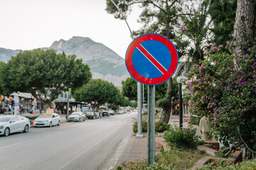 A no parking traffic sign on a street lined with trees, cars, and shops, with mountains in the background, taken in a scenic town or tourist area.