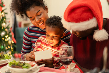 A joyful African American family support their son to open a glowing gift box on Christmas season at dinner table. Moment of magic, love, and surprise on holiday party. Love and bonding people