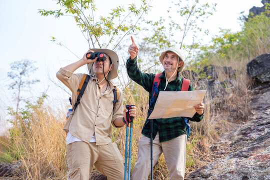 bonding father and son hiking on the mountain together,senior man with backpack holding paper map smiling and looking forward,with adult son carry backpack holding binoculars looking something