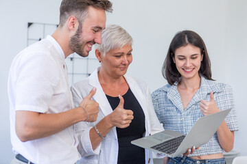 Smiling team of diverse professionals giving thumbs up while looking at laptop, symbolizing business success, team achievement, collaboration, and positive workplace culture.