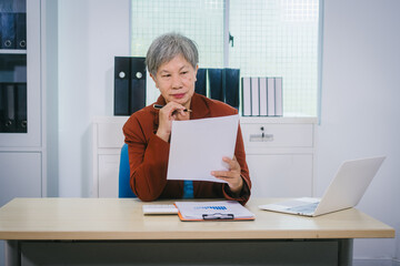 Senior Asian woman with professional office experience, focusing on reviewing financial reports and investment documents, reflecting her expertise in business, finance, and investment strategy