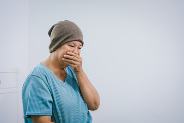 An elderly Asian woman wearing a gray-brown beanie, a light blue shirt, and dark pants sits quietly...