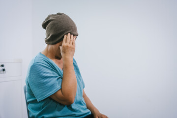 An elderly Asian woman wearing a gray-brown beanie, a light blue shirt, and dark pants sits quietly...