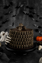 Halloween chocolate cake decorated with chocolate skulls and a skeleton hand on a black cake stand. Spooky ghost, pumpkin, and bat decorations create a dark festive atmosphere.