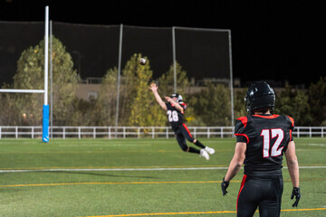 American football player in black jersey jumping for a catch at night on a green field with goalpost and teammate watching