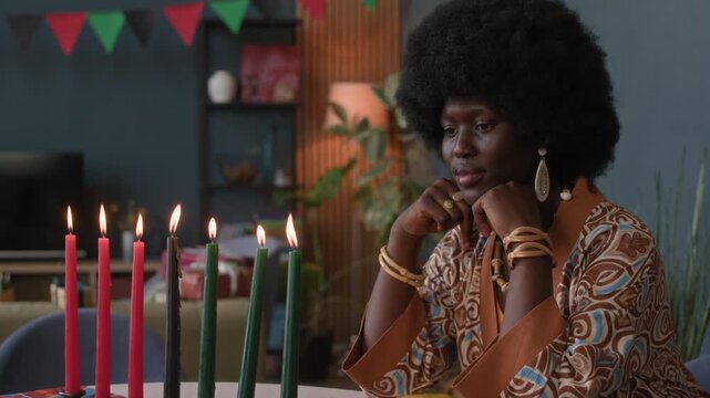 Waist up shot of young African American woman sitting at festive table with patterned cloth, wrapped gifts, glowing candles, and vegetables, honoring Kwanzaa at home