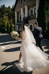 Groom in black suit hugging slim, healthy bride in white dress holding bouquet of white flowers as they walk through the city