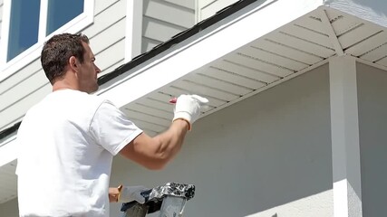 Dedicated man painting house exterior soffit and eaves with a white paintbrush focusing on a home improvement DIY project Professional painter renovating outdoo