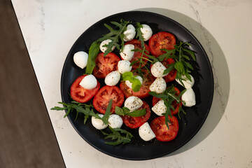 Caprese salad is served on a dark plate. Ripe red tomato slices and mozzarella balls are garnished with fresh arugula and basil leaves on a white surface.