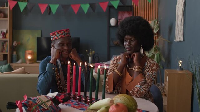 Black couple in traditional clothing sitting at festive table, peacefully admiring lit Kwanzaa candles in bright, cozy living room filled with warmth and cultural celebration