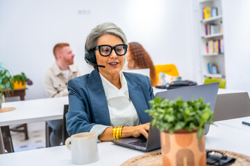 Senior woman working in office wearing headset, using laptop