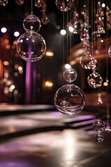 Suspended clear glass orbs of varying sizes hang in a dimly lit interior space, casting soft reflections on a stage-like setting with purple lighting