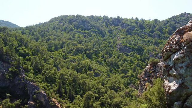 An aerial or scenic view showcasing the vibrant, lush green forested mountains and rocky outcrops under a clear blue sky, capturing the natural beauty of the landscape near Turunc, Turkey.