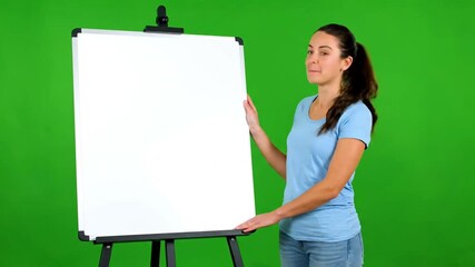 A young woman in a blue t-shirt stands next to a blank white flip chart on an easel, presenting against a solid green screen background. - Powered by Adobe