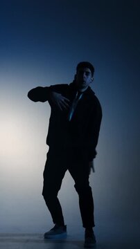 A man in a hoodie performs modern street dance as a graphic silhouette against a blue smoky gradient in a minimalist studio, filmed in slow motion.