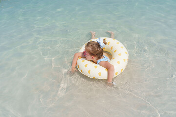 Little blond girl with rubber ring enjoying the blue sea water on the beach 
